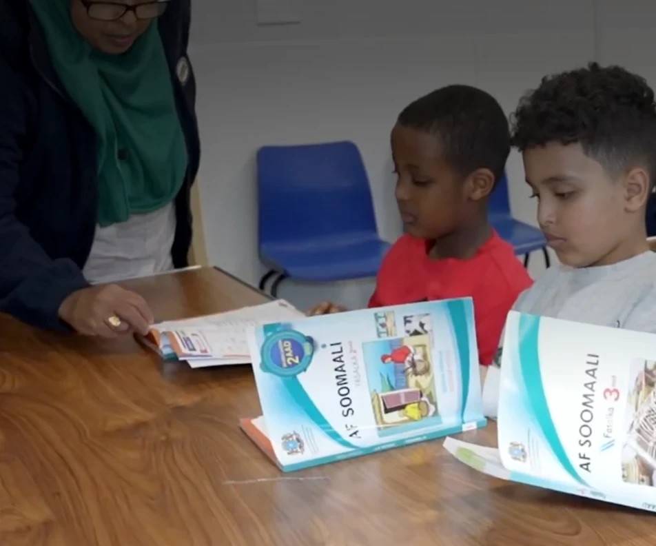 A teacher helping two children read the Af Soomaali textbooks in a Somali Language Centre class